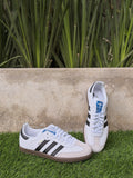 Tenis 𝐋𝐔𝐀𝐋 Adidas Samba blancos de piel con franjas negras, puntera de ante y suela de goma color caramelo.