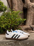 Tenis 𝐋𝐔𝐀𝐋 Adidas Samba blancos con franjas negras, puntera de ante y suela de goma caramelo en un entorno natural.