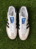 Tenis 𝐋𝐔𝐀𝐋 Adidas Samba blancos de piel con franjas negras, puntera de ante y lengüeta con logo azul sobre pasto.