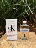 Perfume unisex 𝐋𝐔𝐀𝐋 CK Everyone de Calvin Klein con frasco de vidrio transparente y caja blanca en base de madera
